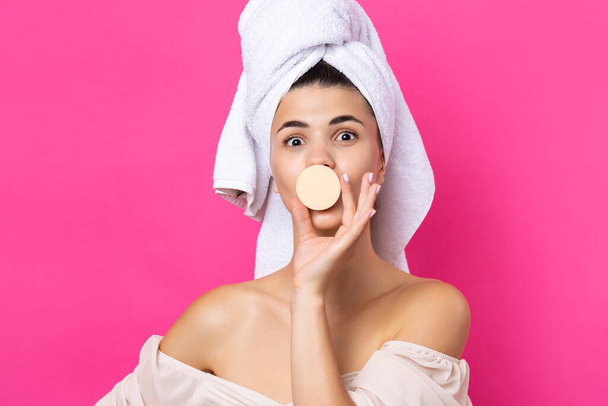 Beautiful cheerful attractive girl with a towel on her head, holds a sponge against a pink background. - 写真・画像