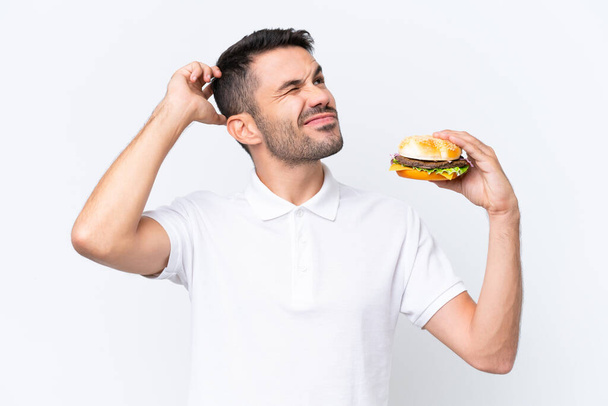Fiatal jóképű kaukázusi férfi tartja a hamburgert az elszigetelt háttér felett, kétségei vannak, és zavaros arckifejezéssel - Fotó, kép