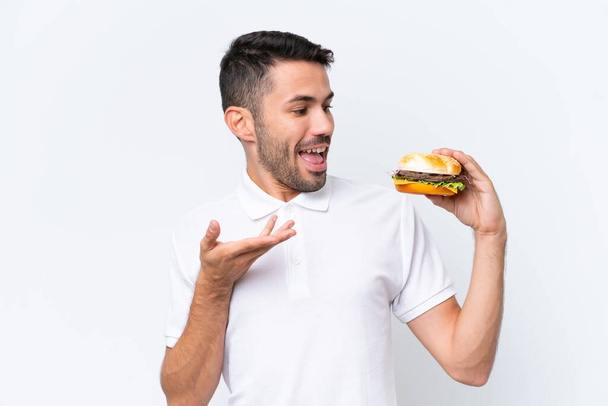 Fiatal jóképű kaukázusi férfi kezében egy burger felett elszigetelt háttér meglepetés arckifejezés - Fotó, kép