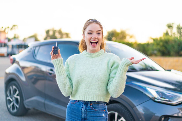 Młoda ładna dziewczyna trzyma kluczyki do samochodu na zewnątrz z wstrząśniętym wyrazem twarzy - Zdjęcie, obraz