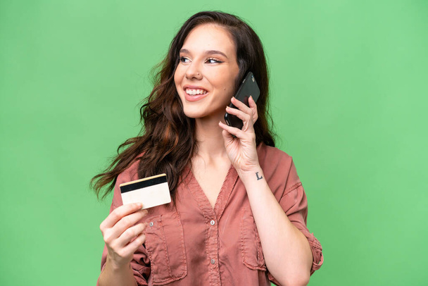 Junge Kaukasierin mit isoliertem Hintergrund führt ein Gespräch mit dem Handy und hält eine Kreditkarte in der Hand - Foto, Bild