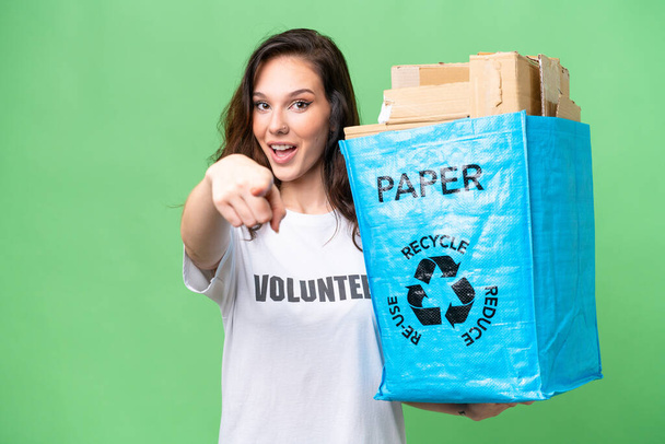 Junge kaukasische Frau hält eine Recyclingtüte voller Papier zum Recyceln über isolierten Hintergrund überrascht und zeigt nach vorne - Foto, Bild