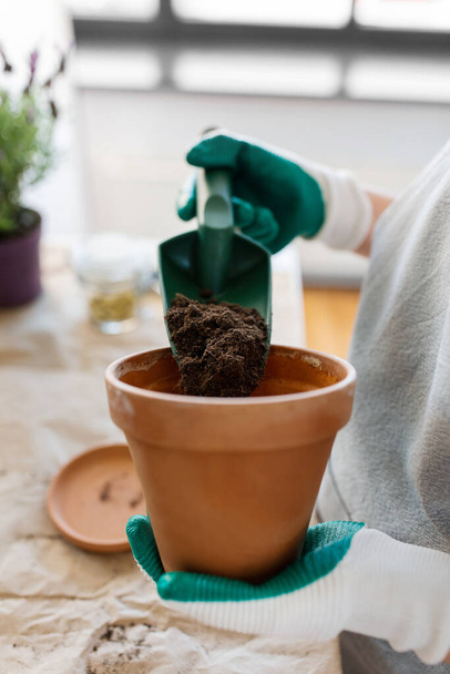 people, gardening and planting concept - close up of woman in gloves with trowel pouring soil to flower pot at home - Photo, image