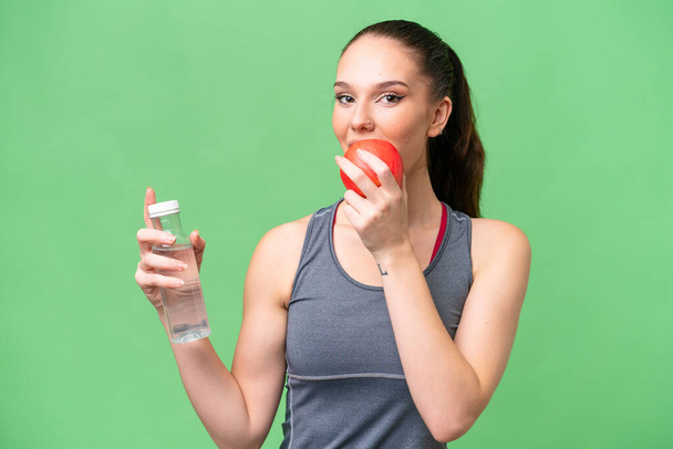 Mujer caucásica joven sobre fondo aislado con una botella de agua y comer una manzana - Foto, Imagen