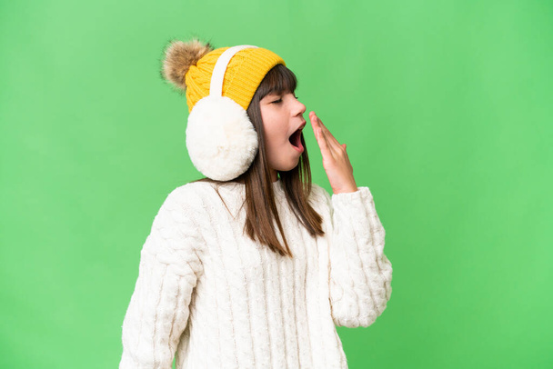 Piccola ragazza caucasica che indossa manicotti invernali su sfondo isolato sbadigliando e coprendo la bocca spalancata con la mano - Foto, immagini