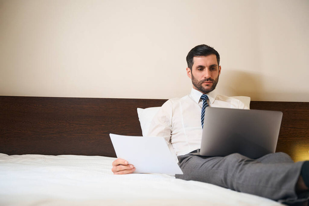 Hombre guapo medio sentado en almohadas, situado en la cama con documentos de trabajo y un ordenador portátil - Foto, imagen
