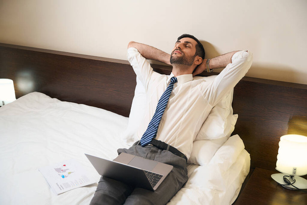 Man closed his eyes from fatigue, he, without undressing, works on a laptop in a comfortable bed - Photo, image