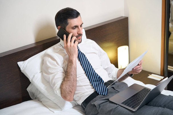 Man in office clothes is studying working documents and talking on the phone, he is located on a large bed - Foto, Imagen