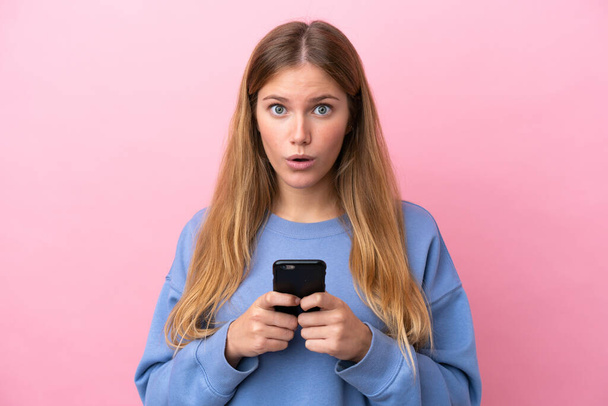 Young blonde woman isolated on pink background looking at the camera while using the mobile with surprised expression - Photo, Image