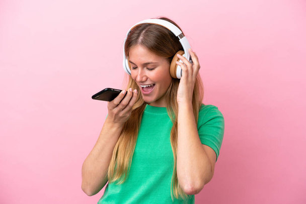 Junge blonde Frau isoliert auf rosa Hintergrund Musik hören mit einem Handy und singen - Foto, Bild