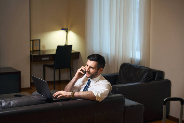 Male in business clothes works in the relaxation area of hotel room, he uses laptop and phone - Foto, Imagen