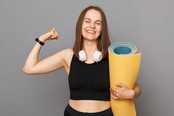 Satisfied delighted girl holding fitness mat in her hands posing isolated over gray background showing her biceps and triceps after athletic workout. - Foto, imagen