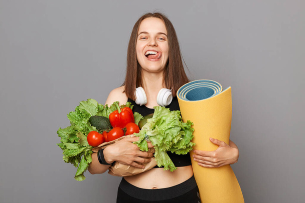 Entzückte Kaukasierin mit Bio-Gemüse und Karemat steht isoliert vor grauem Hintergrund und zeigt hungrige Zunge nach hartem Sport-Training. - Foto, Bild