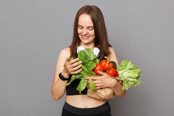 Vonzó mosolygós kaukázusi nő kezében bio zöldségek álló elszigetelt szürke háttér nézi zöld saláta élvezi az egészséges táplálkozás. - Fotó, kép