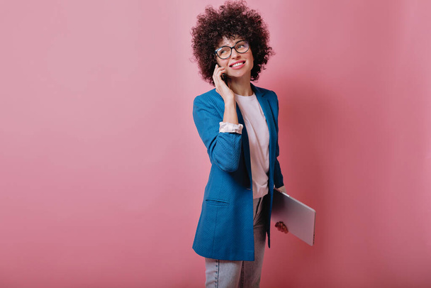 Excited caucasian girl with curly hairstyle posing with smartphone on pink background. Indoor portrait of amazed stylish woman in casual attire holding cell and laptop - Fotó, kép