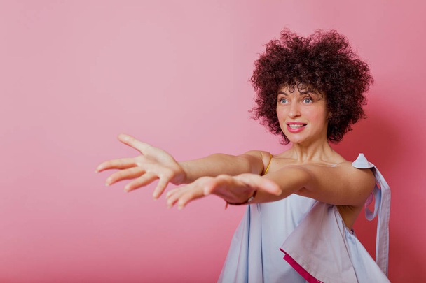 Studioporträt einer jungen schönen Frau in farbenfroher lässiger Kleidung, die fröhlich ist und die Hände nach vorne hebt und auf rosa Hintergrund lächelt. Menschen-Porträt-Konzept - Foto, Bild