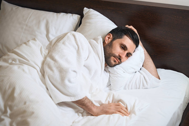 Fáradt szállodai vendég fekszik az ágyban puha párnák, ő egy fürdőköpeny - Fotó, kép