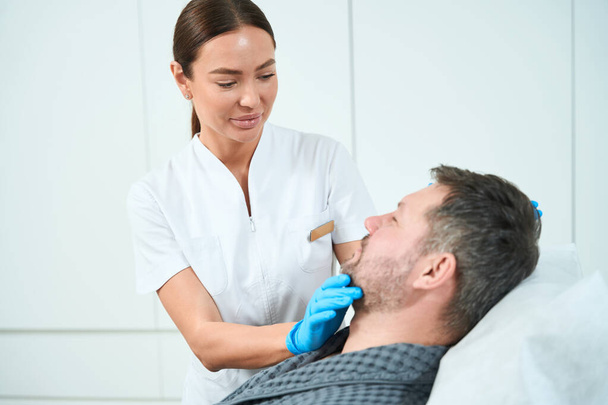 Cosmetologista profissional examina o rosto de um paciente de meia-idade, um homem confortavelmente assentado em um sofá cosmetologia - Foto, Imagem