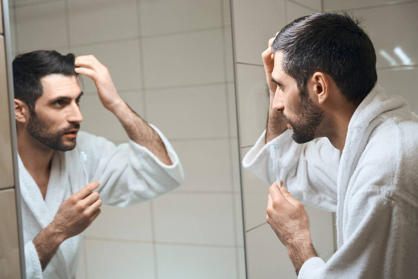Unshaven male stands in front of a mirror in the bathroom, he looks at his reflection - Zdjęcie, obraz