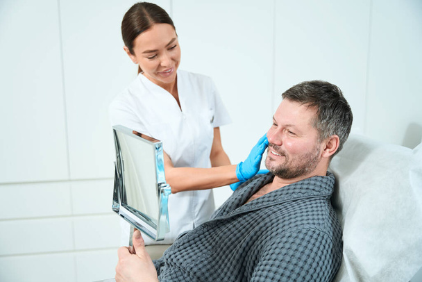 Smiling man sits in front of a mirror at a consultation with a beautician, a specialist works in protective gloves - Photo, Image