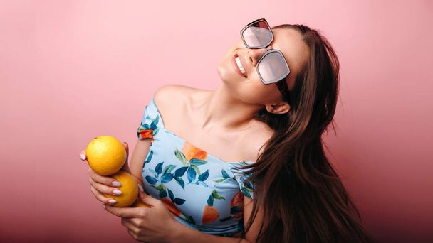 Young smiling woman in sunglasses having fun holding oranges isolated over pink background. People emotions concept  - Фото, изображение