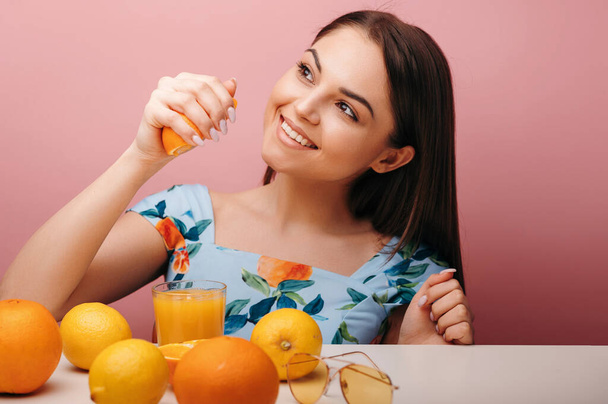 Brunette woman preparing orange juice and holding fruits at the table. Isolated studio portrait. Healthy lifestyle concept  - Fotó, kép