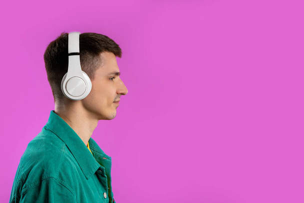 Positive young man listening music, enjoying dance with headphones on violet studio background. Radio, wireless modern sound technology, online player. High quality - Valokuva, kuva