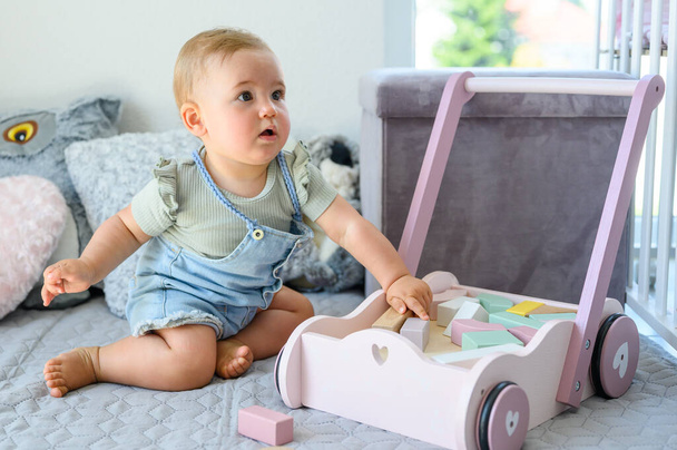 Full body of cute little baby in casual clothes sitting on bed while playing with wooden blocks in toy cart in bedroom at home - Φωτογραφία, εικόνα