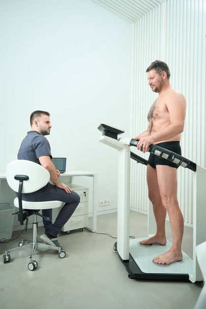 Patient with a naked torso at the diagnostic procedure of bioimpedancemetry, next to an employee in a medical uniform - Fotografie, Obrázek