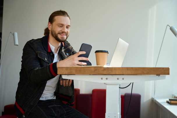 Smiling man in stylish clothes browsing cell phone while sitting in modern room - Fotoğraf, Görsel