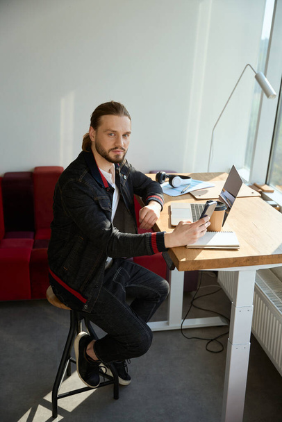 Komea mies katselee kameraa matkapuhelinta käyttäessään ja istuu tuolilla coworking-tilassa - Valokuva, kuva