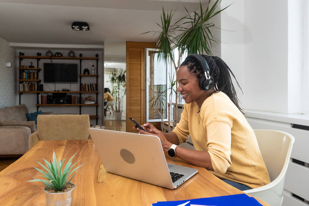 Start up businesswoman, African American woman wearing headphones music and holding mobile phone using bluetooth listening music while sitting resting after work happy and relaxing at home office - Fotografie, Obrázek