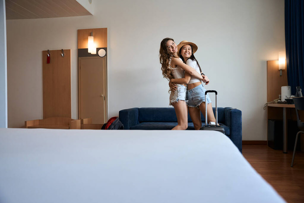 Sorrindo amigos do sexo feminino de pé perto do sofá e abraçando depois de verificar no albergue - Foto, Imagem
