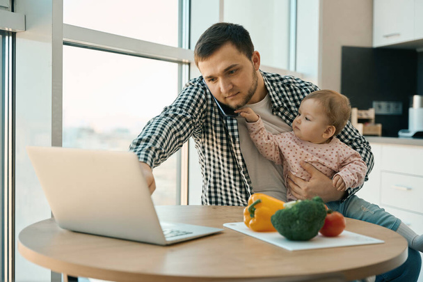 Bearded man works from home with child in his arms, laptop and a variety of vegetables are on the table - Foto, Imagem