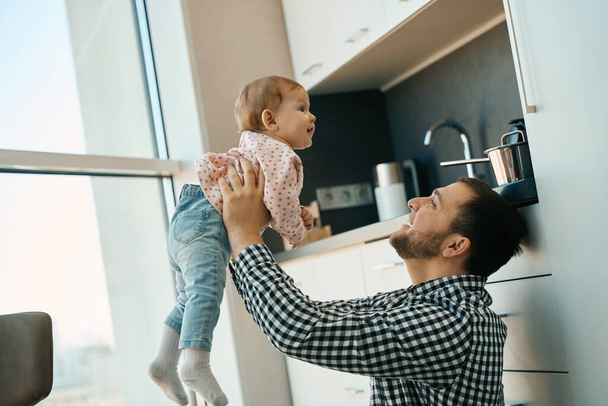 Bearded father joyfully plays with a pretty little daughter, the girl is wearing cute baby clothes - Φωτογραφία, εικόνα