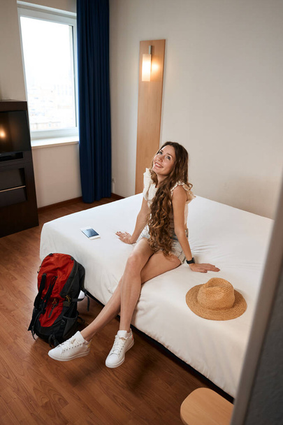 Улыбающаяся женщина сидит на кровати рядом с рюкзаком и смотрит вверх в общежитии - Фото, изображение