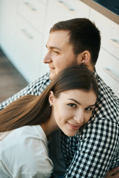 Beautiful man and woman are sitting, gently embracing, on the floor in the kitchen, guys in simple home clothes - Foto, imagen