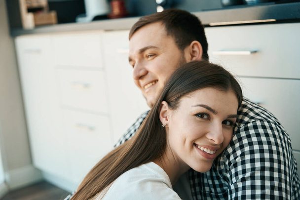 Happy man and woman are sitting, gently embracing, on the floor in the kitchen, male in a plaid shirt - Fotografie, Obrázek