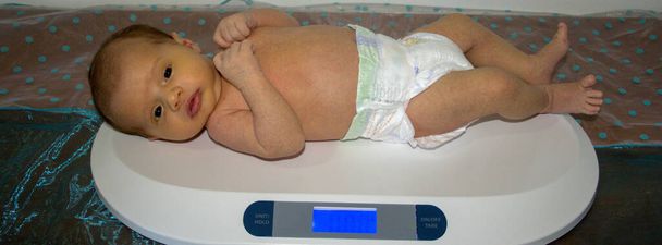 Afbeelding van een pasgeboren baby met een luier terwijl hij op een weegschaal wordt gewogen. Meting van gewicht en groei van pasgeborenen. Horizontale banner  - Foto, afbeelding