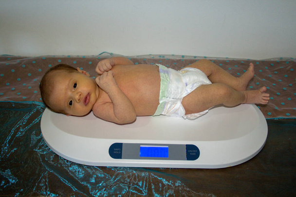 Afbeelding van een pasgeboren baby met een luier terwijl hij op een weegschaal wordt gewogen. Meting van gewicht en groei van pasgeborenen - Foto, afbeelding