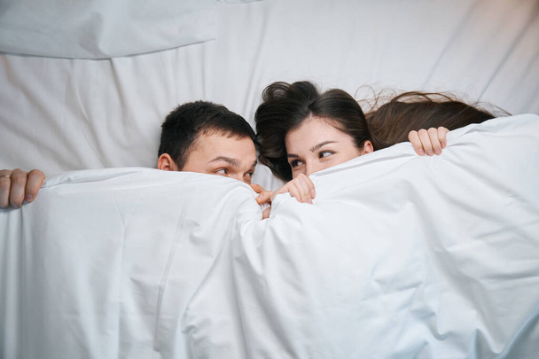Cute husband and wife are basking under the covers on a large bed, they look at each other funny from under the covers - Foto, Imagem