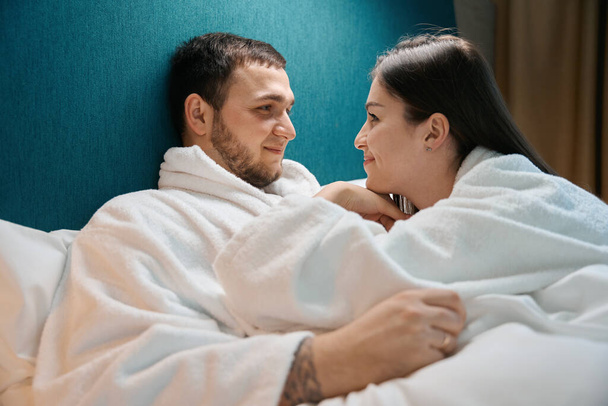 Smiling charming woman looks tenderly at her husband, the spouses bask under the covers on a large bed - Fotoğraf, Görsel