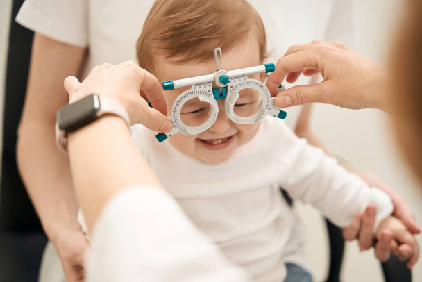 Жіночі руки-окулістки наносять офтальмологічну пробну рамку на радісну дитину в присутності батьків
 - Фото, зображення