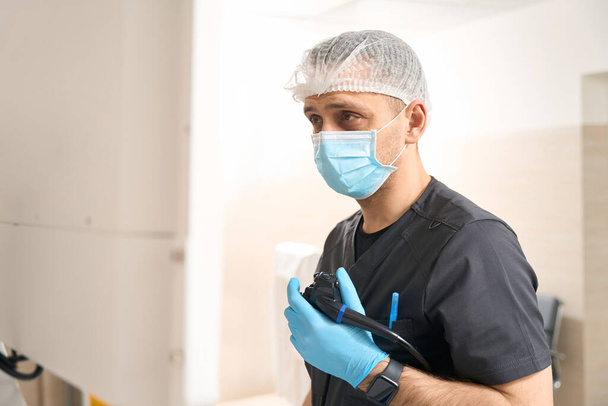 Waist-up portrait of diagnostician holding endoscope with gloved hand while staring at monitor screen during endoscopy - Photo, Image