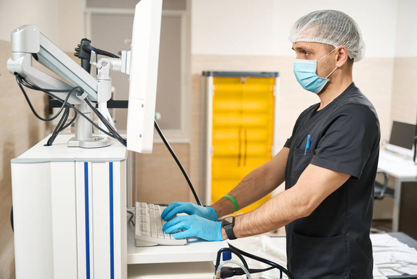 Oldalnézet súlyos diagnosztikus orvosi maszk és eldobható kesztyű gépelés a számítógép billentyűzetén, miközben bámulja monitor képernyő - Fotó, kép