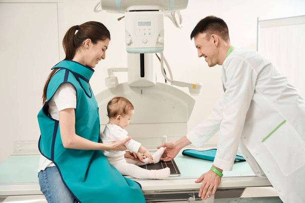 Uśmiechnięty radiograf umieszczający nogę dziecka na stole rentgenowskim w obecności radosnego rodzica płci żeńskiej - Zdjęcie, obraz