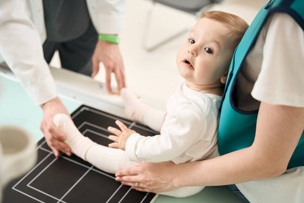 Przycięte zdjęcie radiologa pozycjonującego nogę dziecka na stole radiograficznym wspomaganego przez rodzica płci żeńskiej - Zdjęcie, obraz