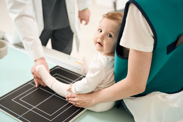 Radyolojik teknoloji uzmanının, ebeveynin yardımıyla bebek bacağını radyografik masaya yerleştirirken çekilmiş fotoğrafı. - Fotoğraf, Görsel