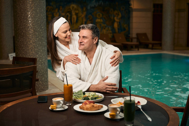 Smiling adult woman in bathrobe hugging from behind pleased mature man at table by swimming pool - Photo, Image