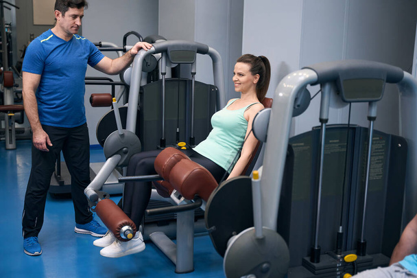 Χαρούμενη ώριμη κυρία εκτελεί καθιστή επέκταση πόδι σε μηχάνημα άσκησης υπό την καθοδήγηση του γυμναστή - Φωτογραφία, εικόνα